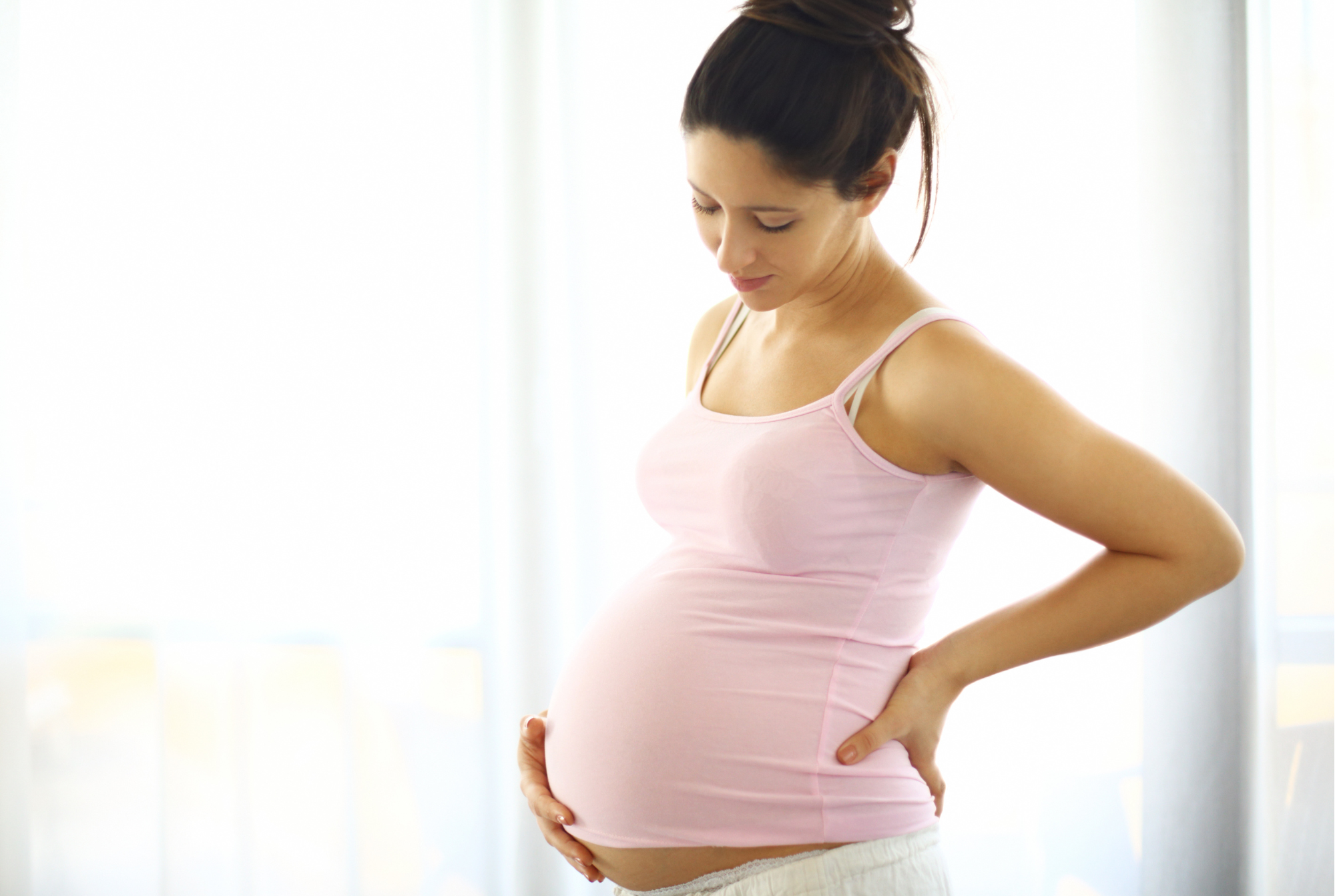 The image depicts a pregnant woman in a pink strappy top and white trousers looking down and holding her bump.