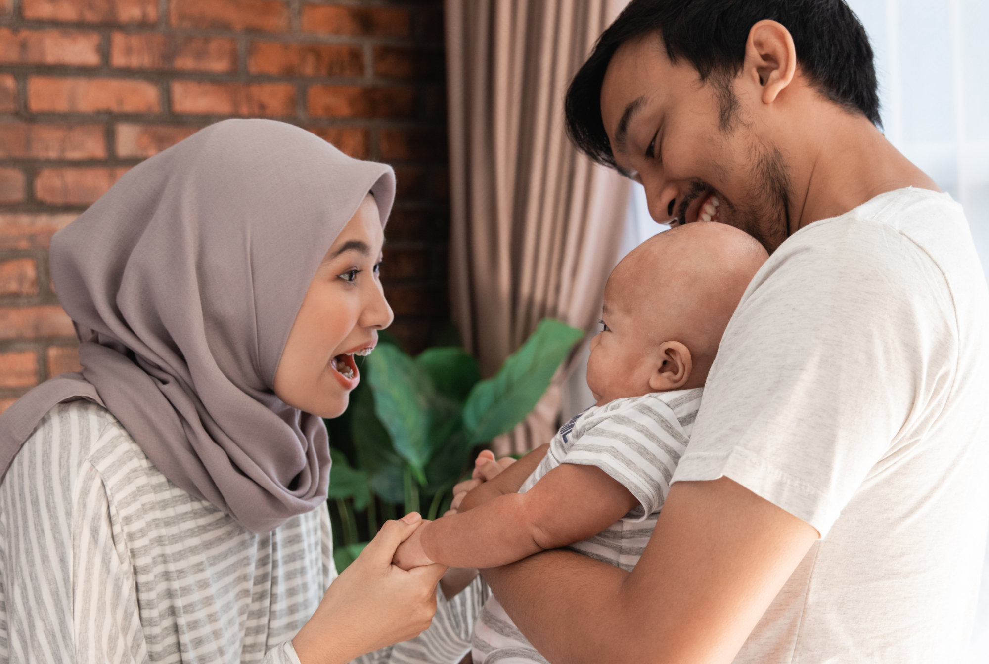 Image depicts a Muslim woman smiling at her baby. The baby is being held by the dad.